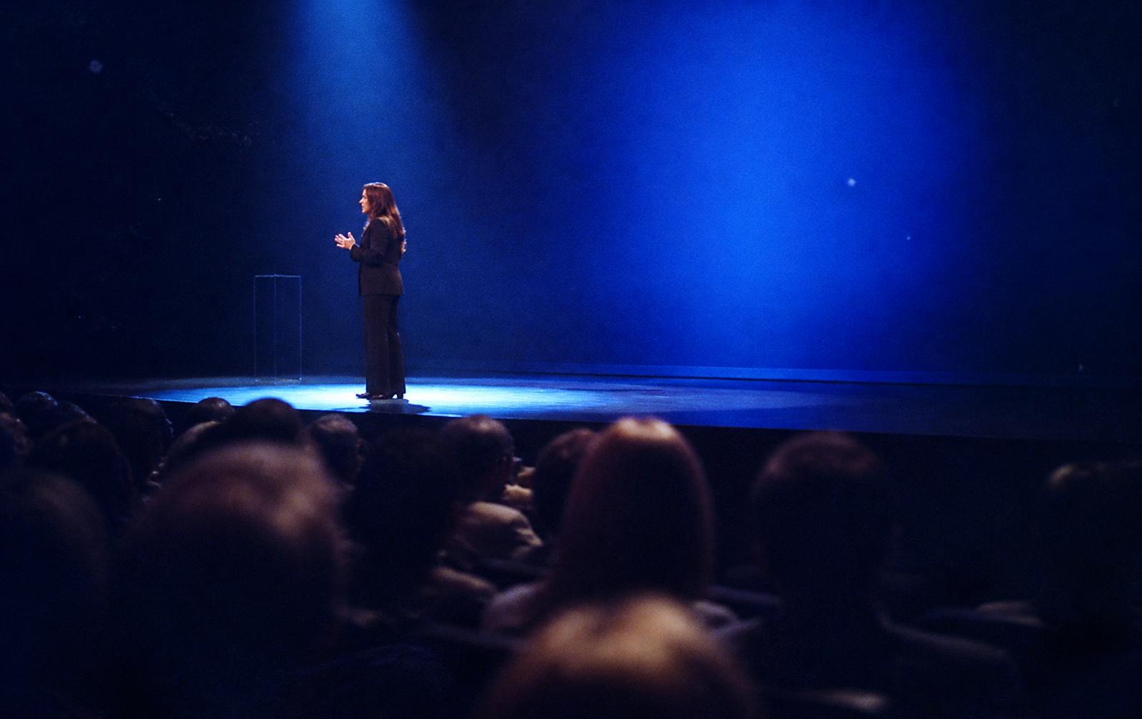 Een vrouw die spreekt op een donker podium, verlicht in kobaltblauw